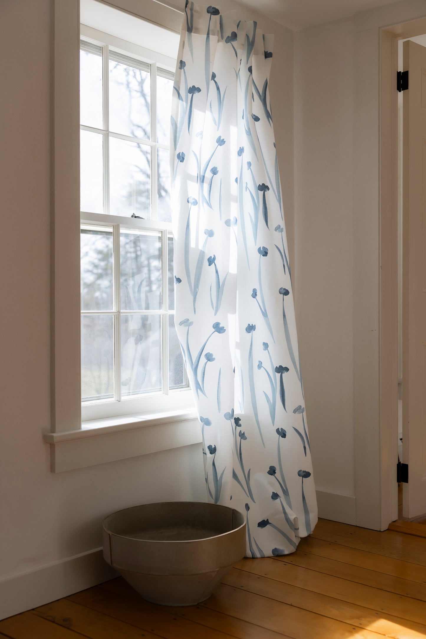 White curtain with blue floral pattern in a room with a window and wooden floor.