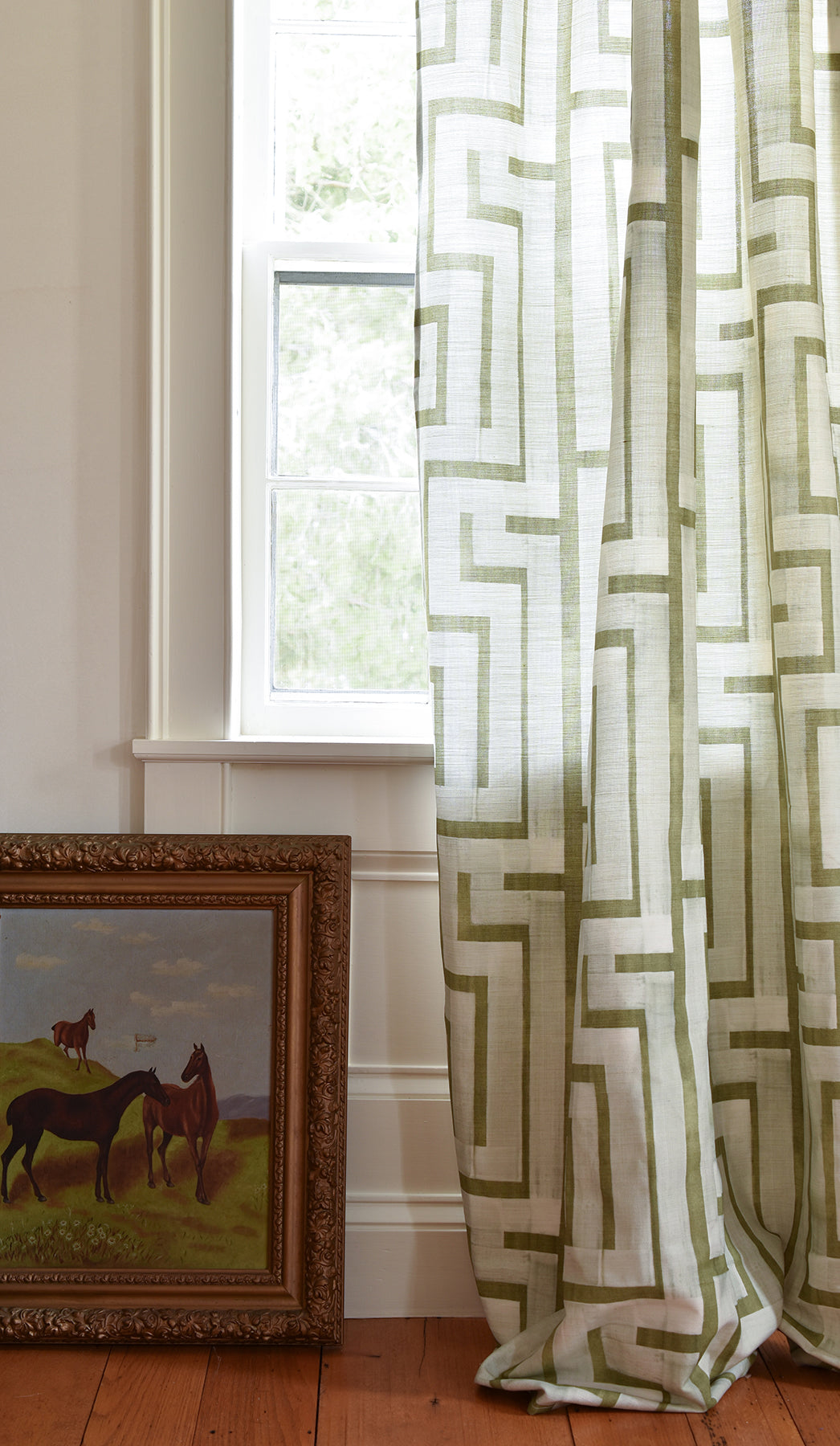 Curtain with geometric pattern next to a window and framed painting of horses.