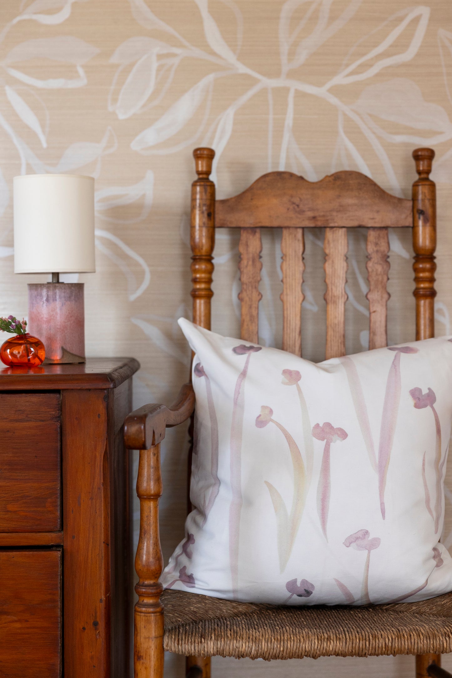 Wooden chair with a decorative pillow against a floral wallpapered wall.