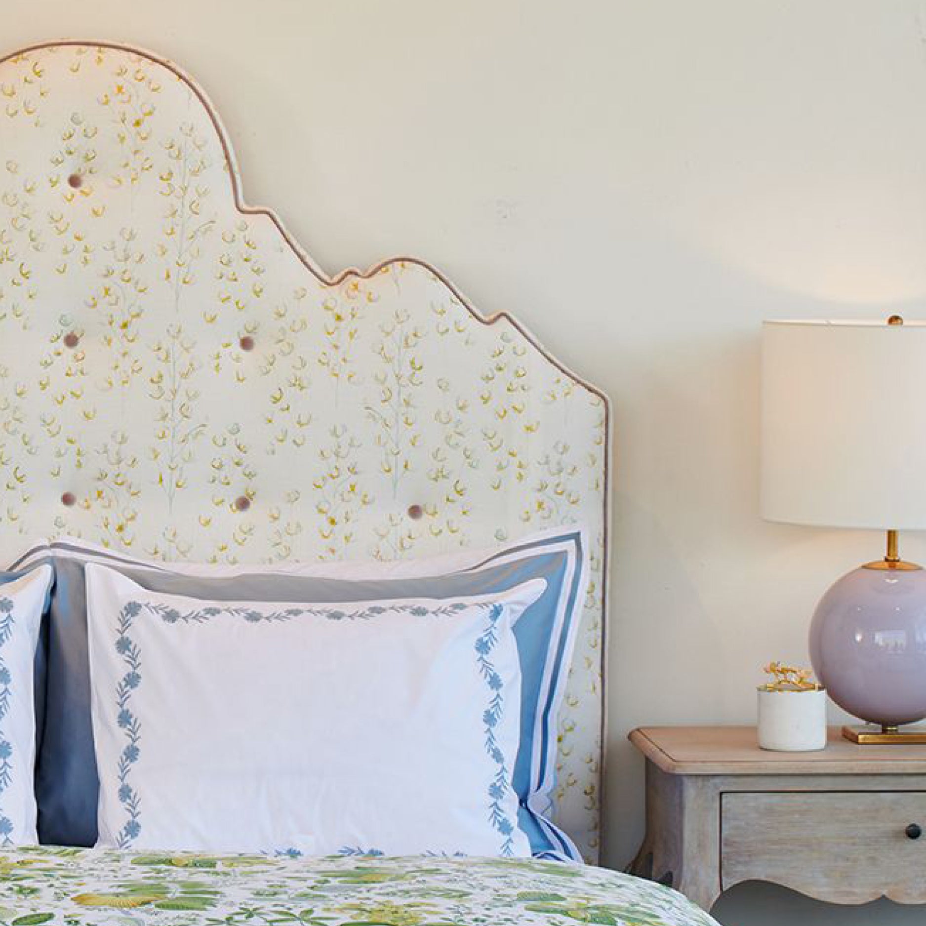 Bedroom with floral headboard, pillows, and nightstand with lamp.