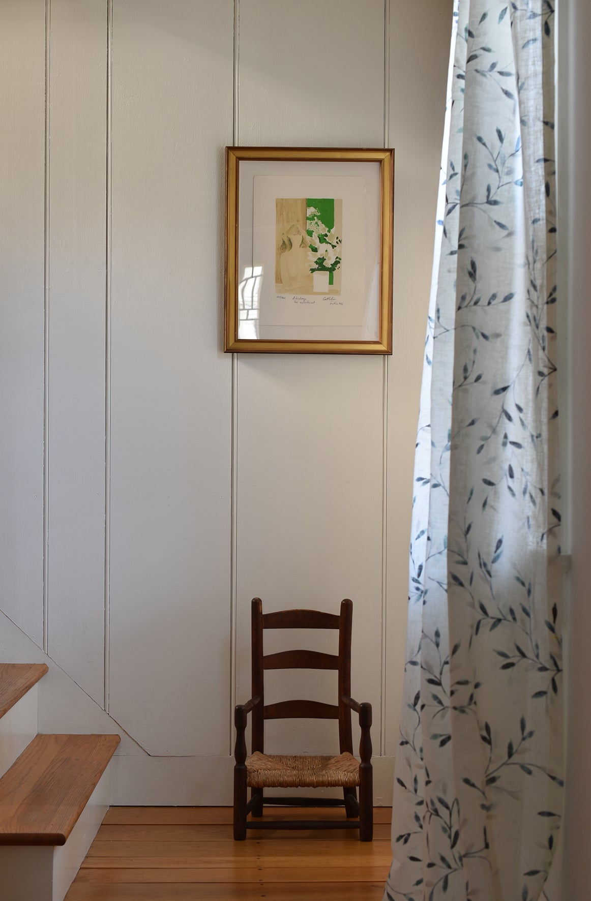 Wooden chair against a white wall with a framed picture, next to a floral-patterned curtain.
