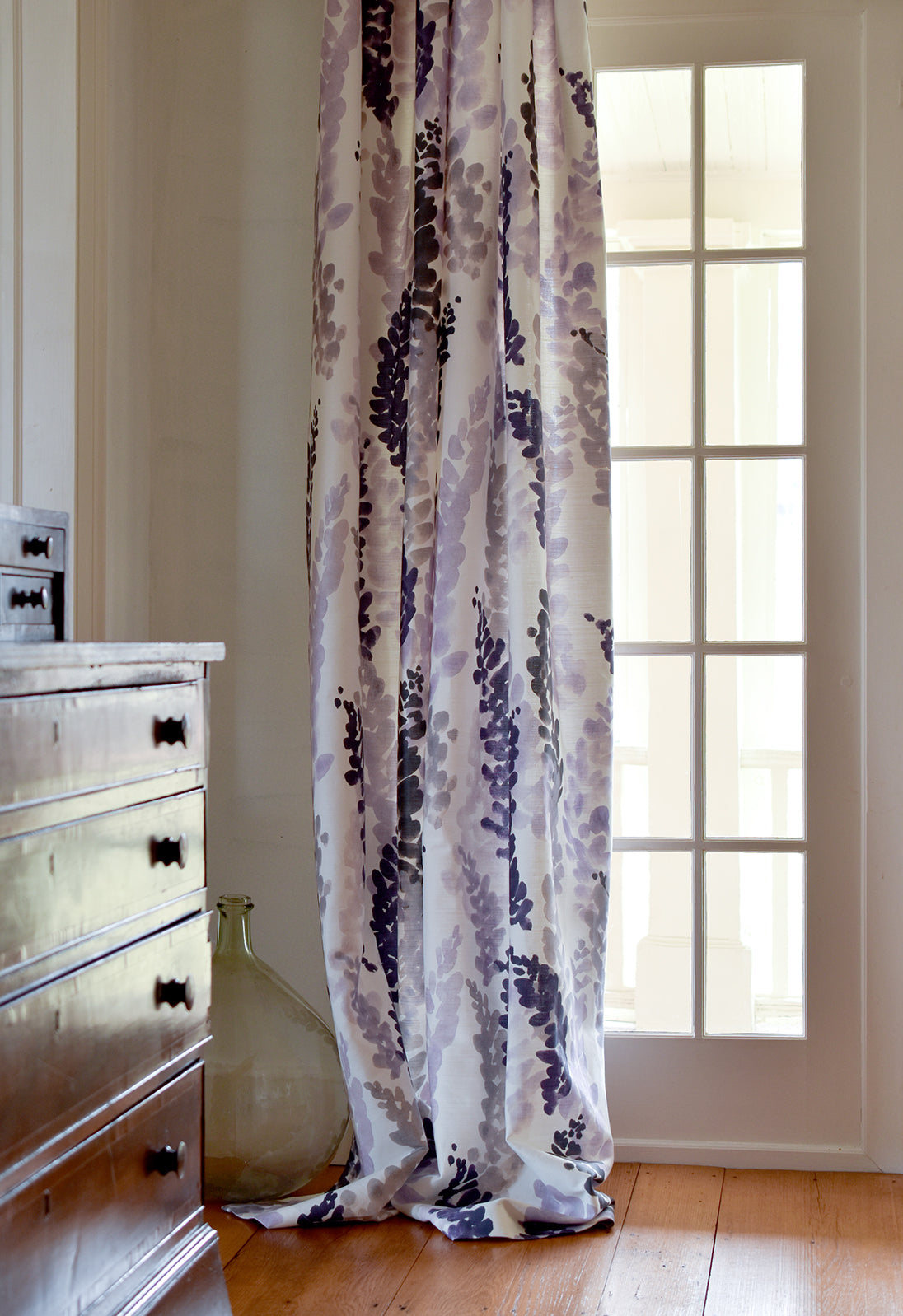 Floral patterned curtain hanging in a room with a wooden floor and a dresser.