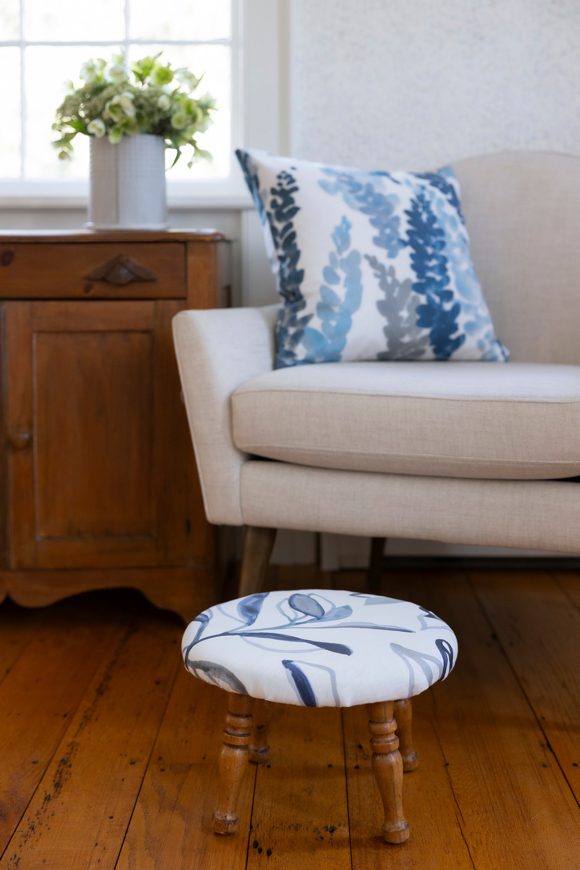 Beige sofa with a decorative pillow and a matching footstool in a room with wooden flooring and a window.