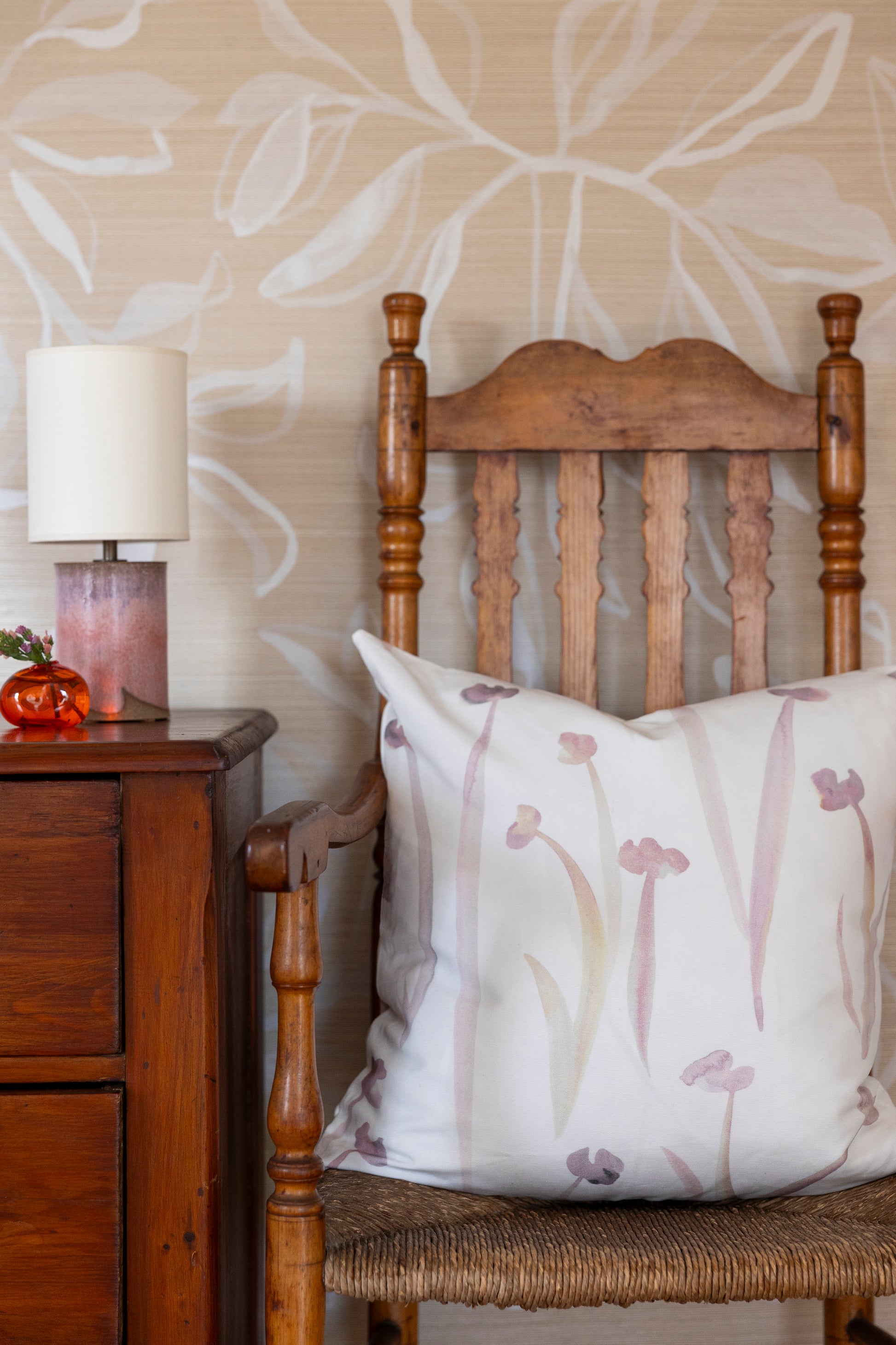 Wooden chair with a decorative pillow against a floral wallpapered wall.